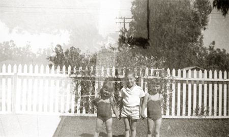 Old black and white summer-time photo of 3 children holding hands in front of white picked fence